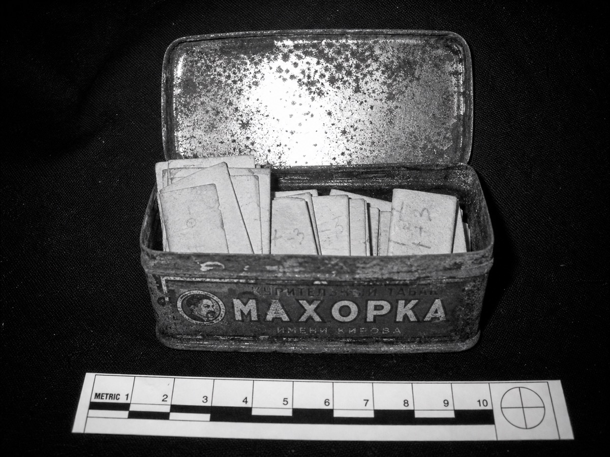 An evidence-style flash photograph of a rusted makhorka tobacco tin, lid open, packed with small folded paper scraps, a measuring scale laid in front.